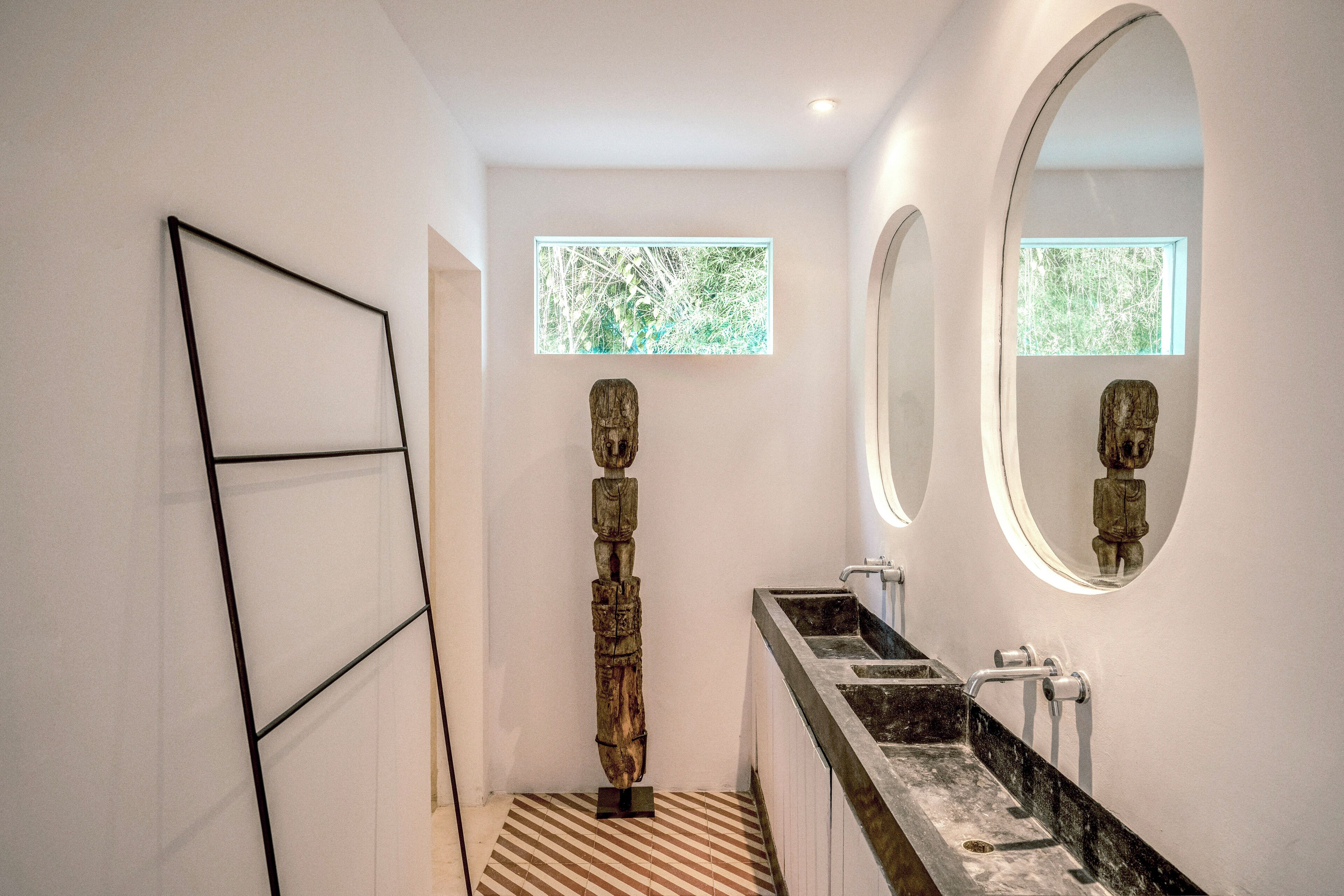 A modern bathroom with a large mirror, a wooden sculpture, and a window with a view of greenery.