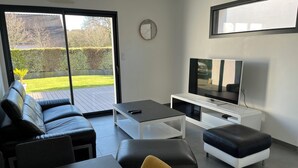 A modern living room with a sofa, ottoman, coffee table, TV, and a sliding glass door leading to a deck. - Maison 4ch/8pers Bourg de Baden, Proche des Plages au Coeur du Golfe du Morbihan (Baden)