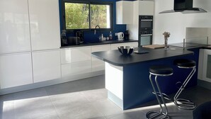 A modern kitchen with white cabinets, a blue accent wall, a large window, and a black range hood. - Maison 4ch/8pers Bourg de Baden, Proche des Plages au Coeur du Golfe du Morbihan (Baden)