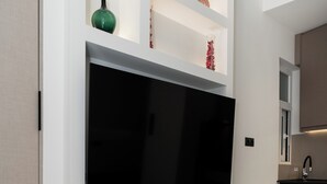 A modern living room with a large flat-screen TV mounted on a white wall, a small seating area with a chair, and a kitchen area visible to the right. - Minimal Studio with Balcony in Central Athens  (Athens)