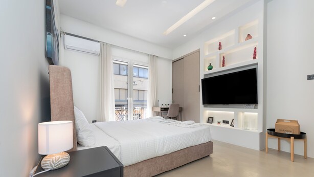 A modern hotel room with a large bed, a TV mounted on the wall, a desk with a lamp, and a view of the city through the window. - Minimal Studio with Balcony in Central Athens  (Athens)