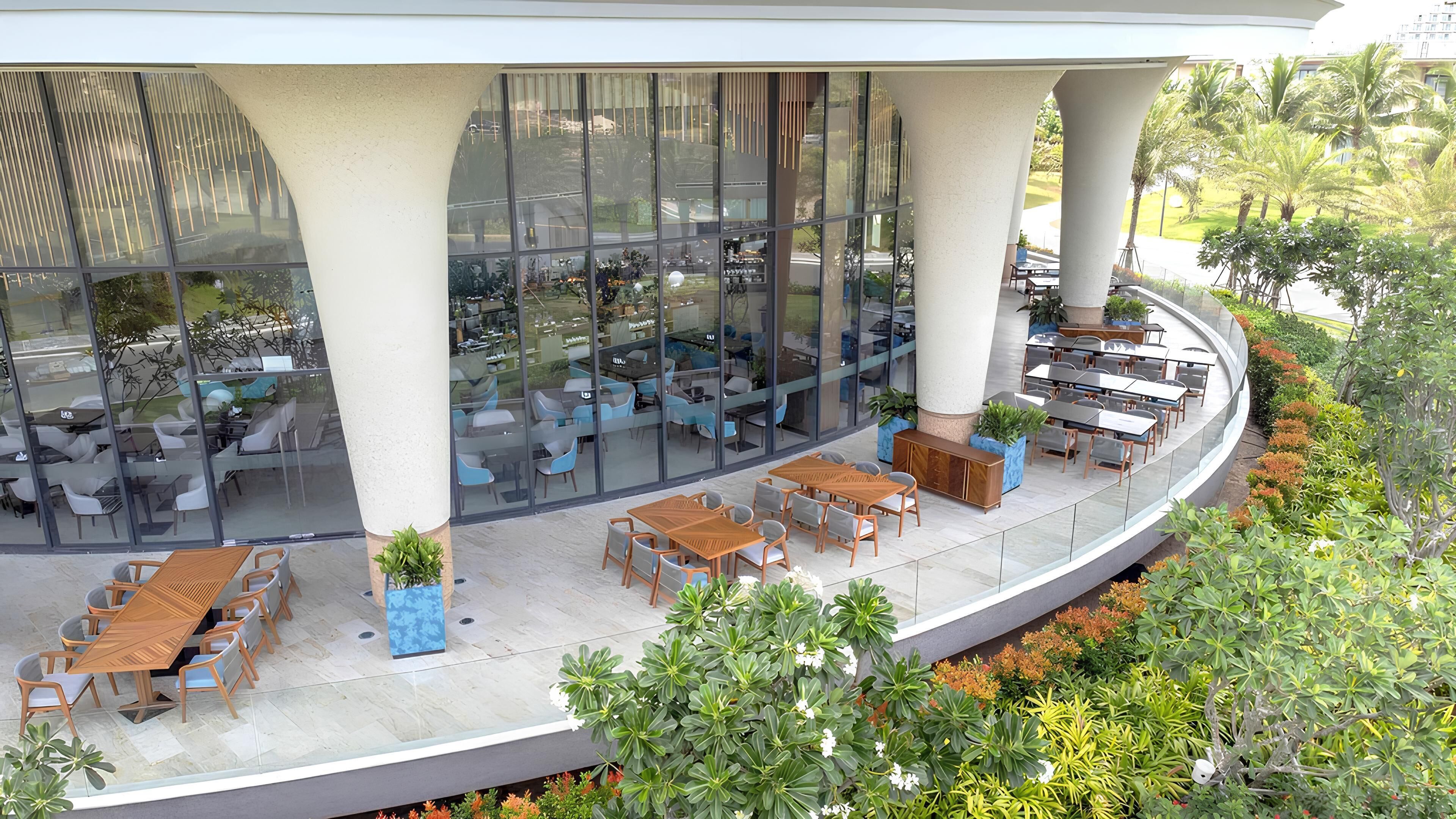 A modern outdoor dining area with multiple tables and chairs, surrounded by glass walls and large pillars.