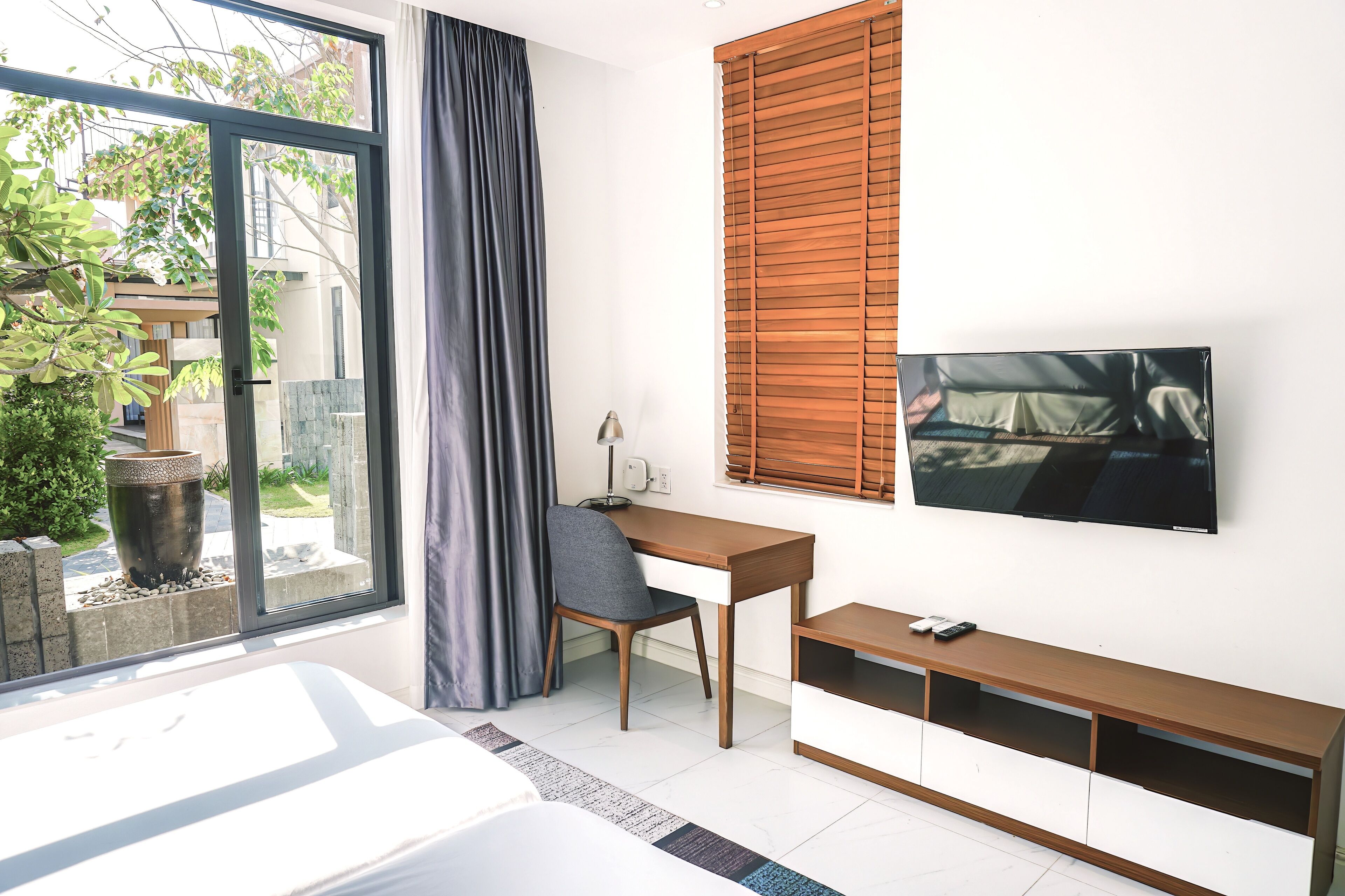 A modern hotel room with a large window, a desk, a chair, a bed, and a TV mounted on the wall.