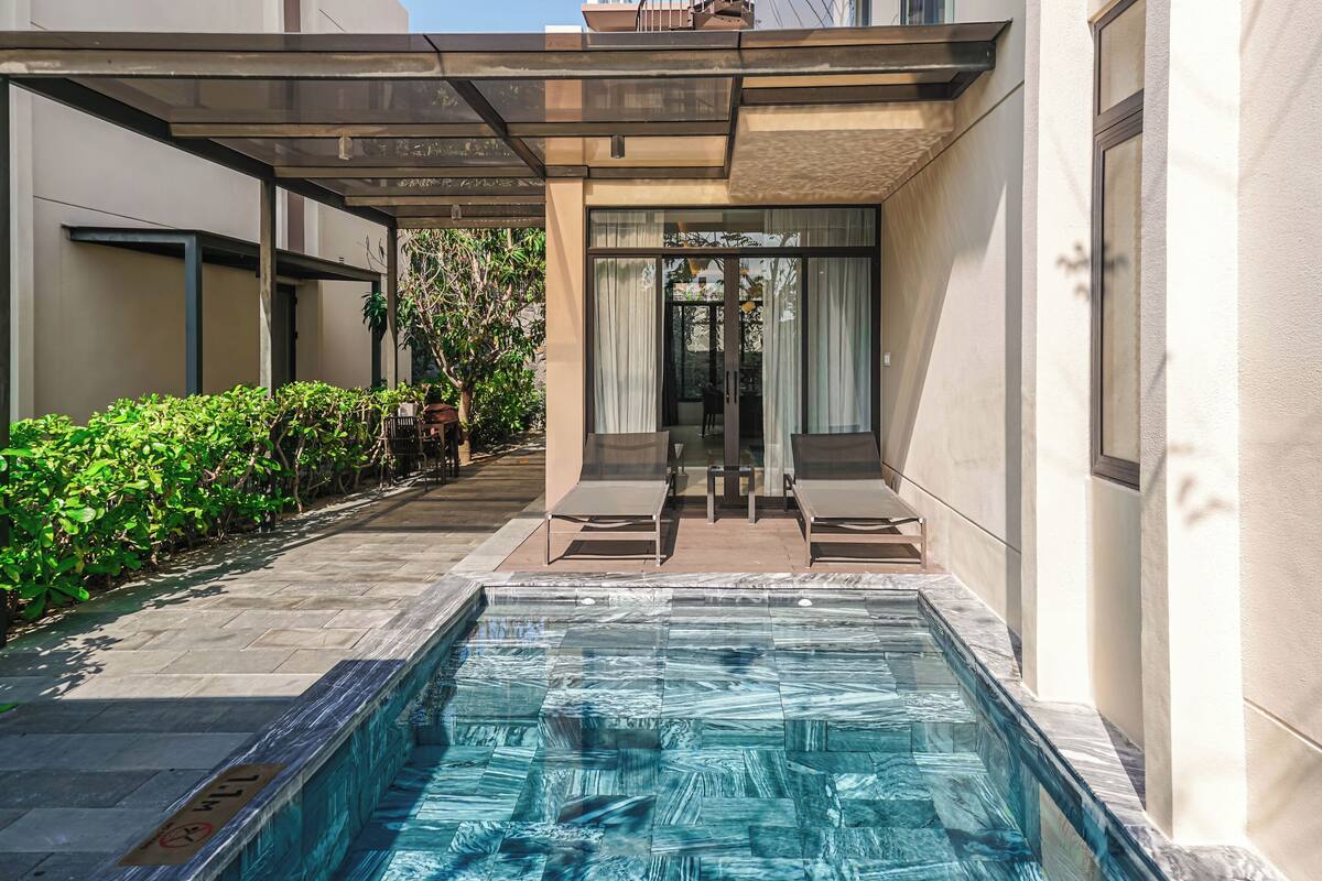 A modern outdoor pool area with a glass enclosure, lounge chairs, and a pathway lined with greenery.