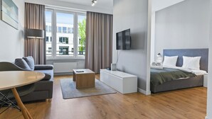 A modern hotel room with a sofa, a small wooden table, a bed with a grey headboard, a TV mounted on the wall, and a window with curtains. - RentPlanet - Apartamenty Sławińska (Warsaw)