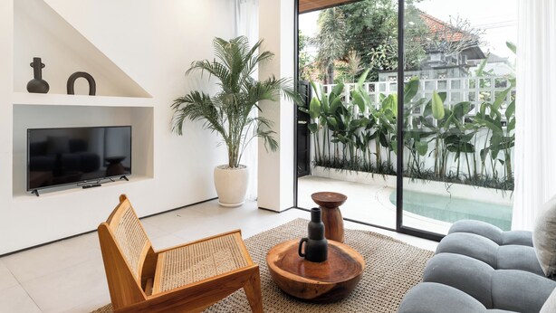 Una sala de estar moderna con televisor, muebles de madera, una planta grande en maceta y vistas a un jardín a través de amplios ventanales.