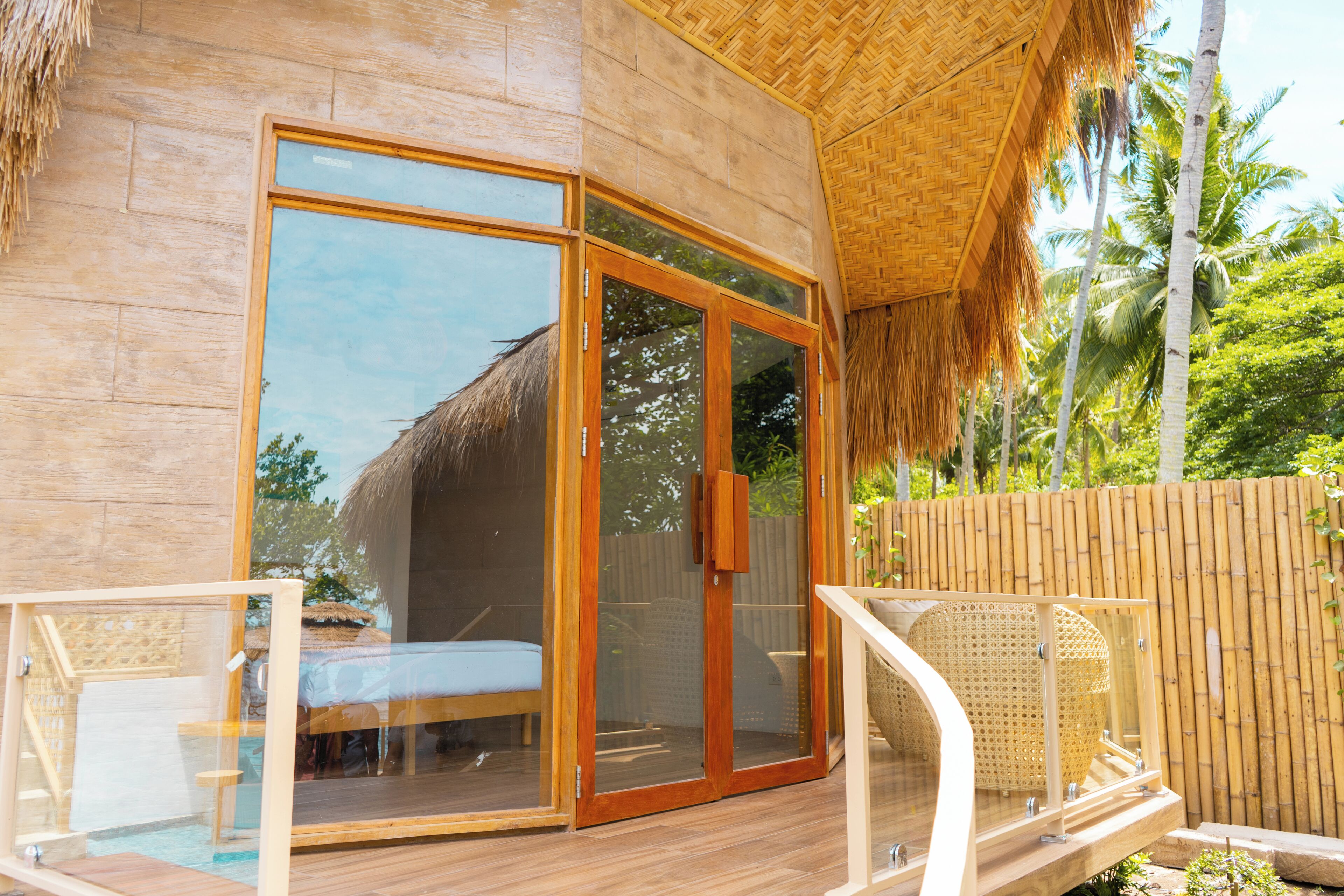 Eine Holzdielen-Terrasse mit einer Glastür, die zu einem Zimmer mit einem Bett und einem Strohdach führt.