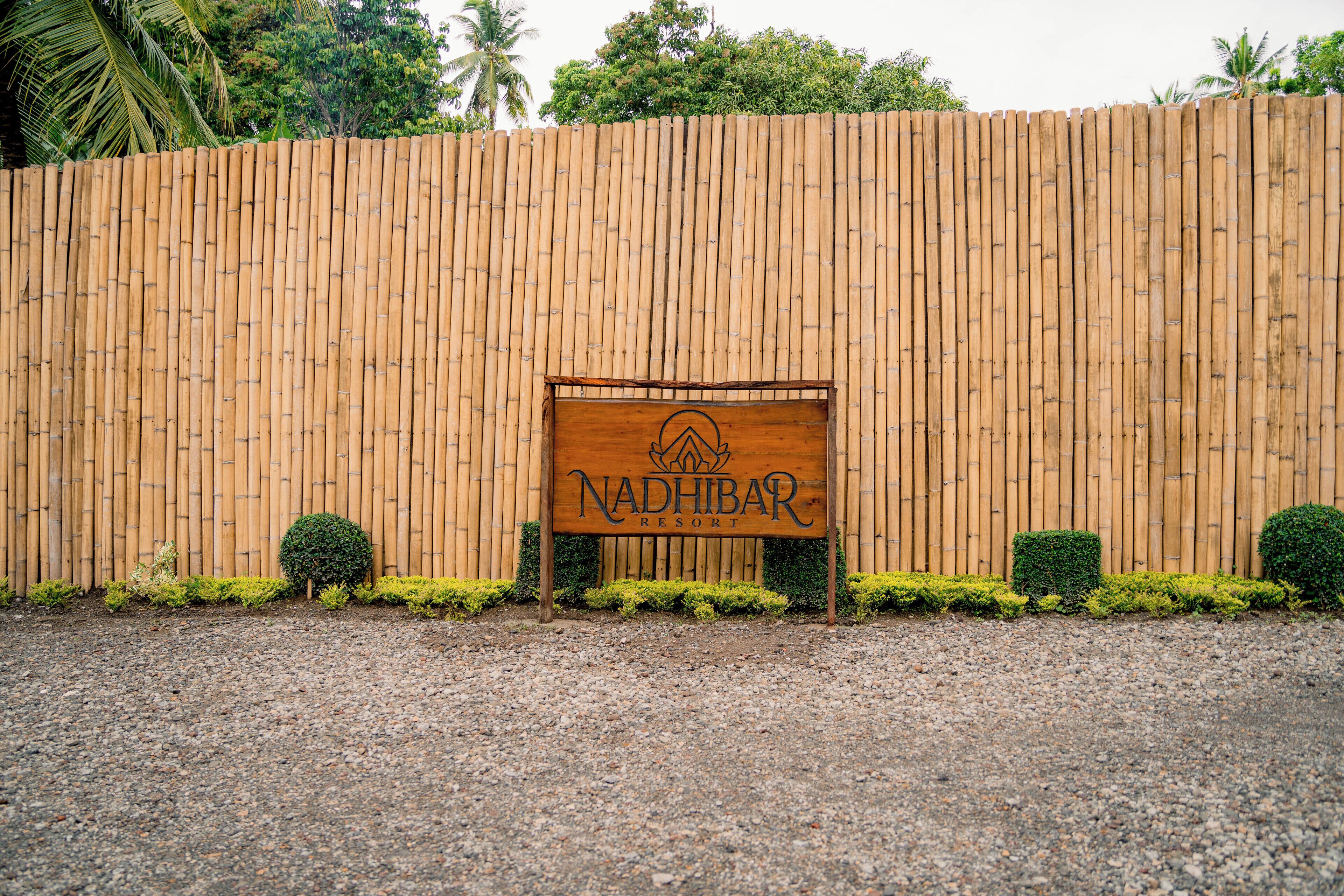 A wooden sign with the text "NADHIBAR RESORT" in front of a bamboo fence.