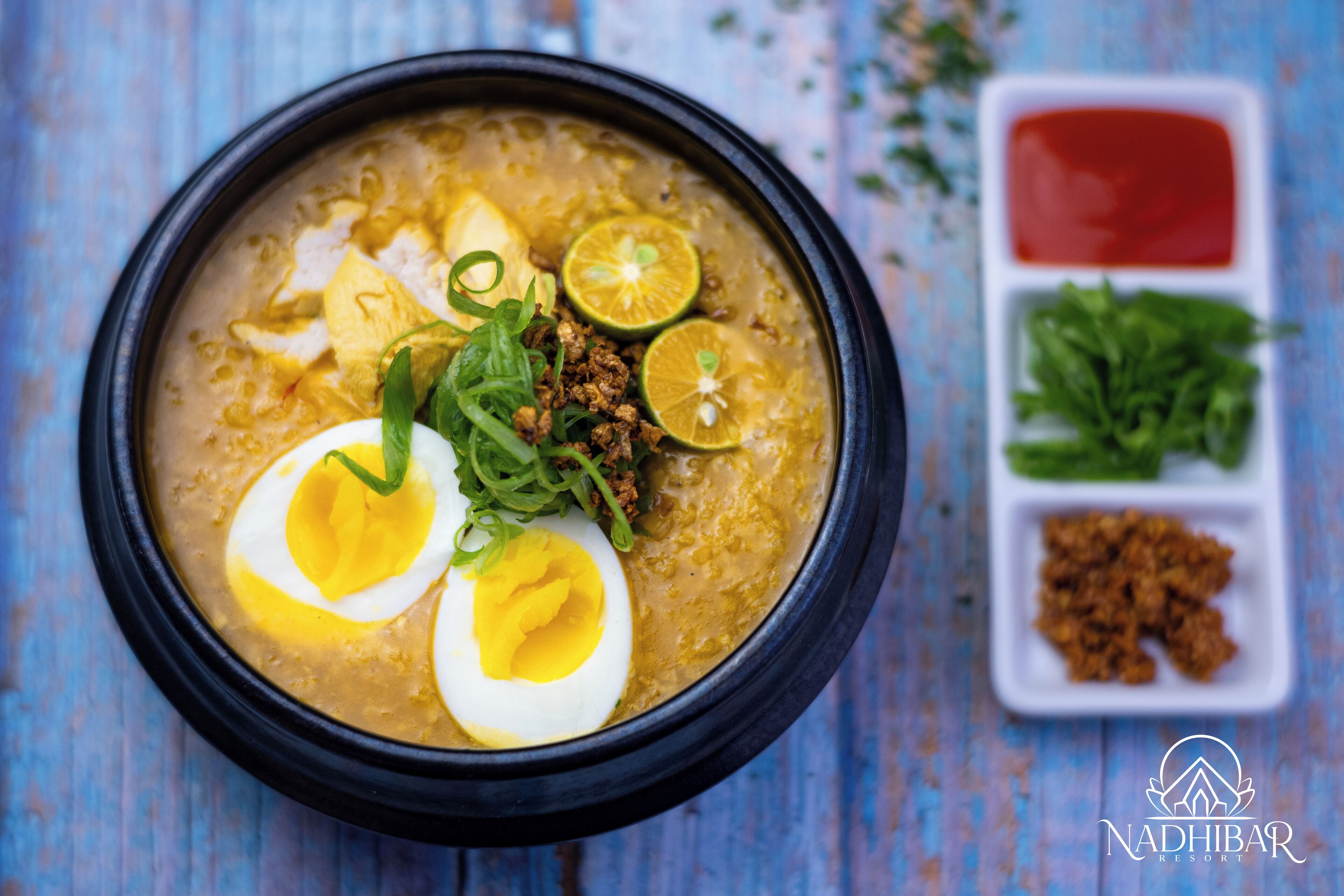 A bowl of soup with boiled eggs, garnished with green onions and lime slices, accompanied by a side of red sauce and greens.