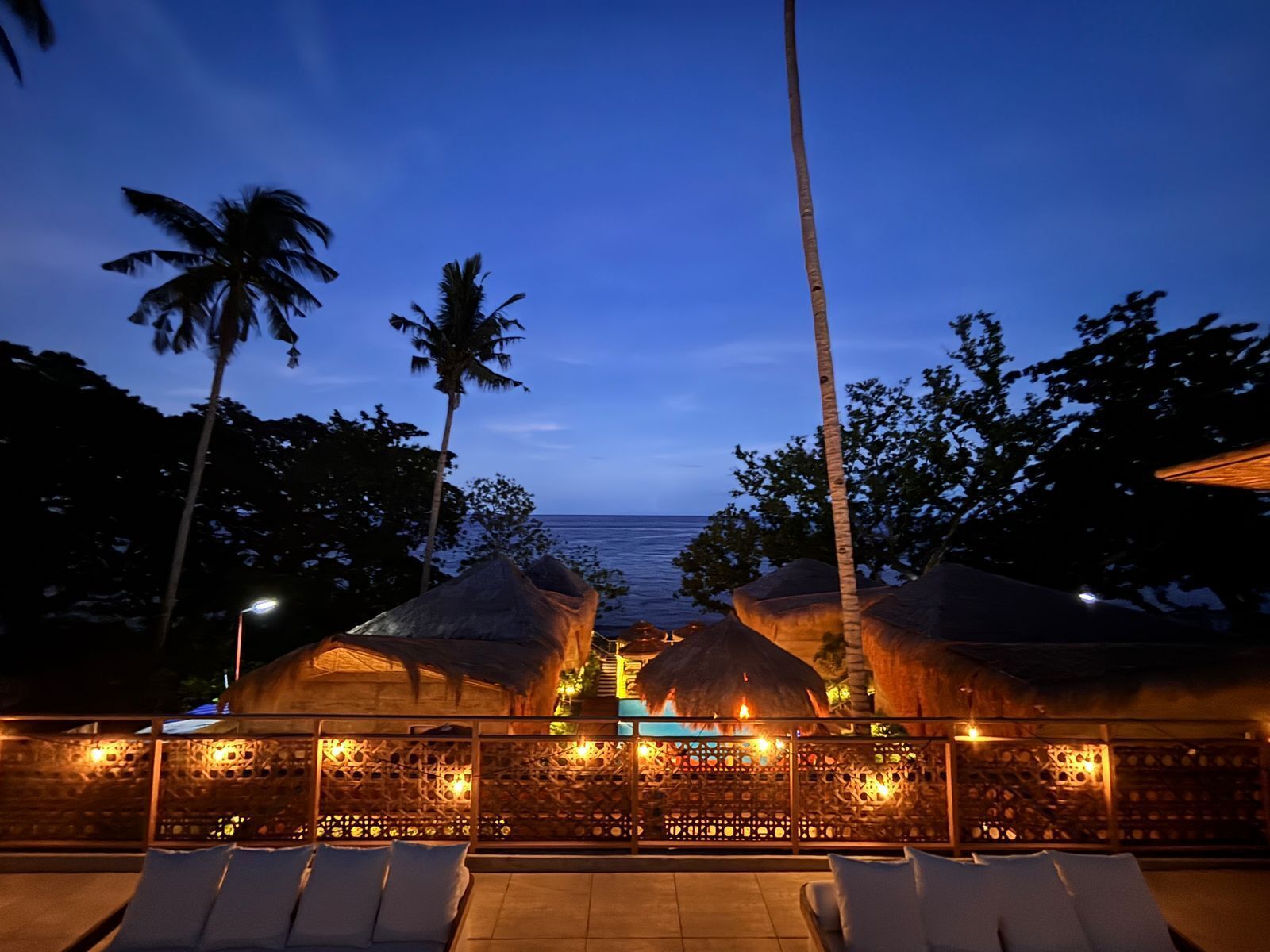 A beachfront resort with thatched-roof structures, lounge chairs, and a view of the ocean at dusk.