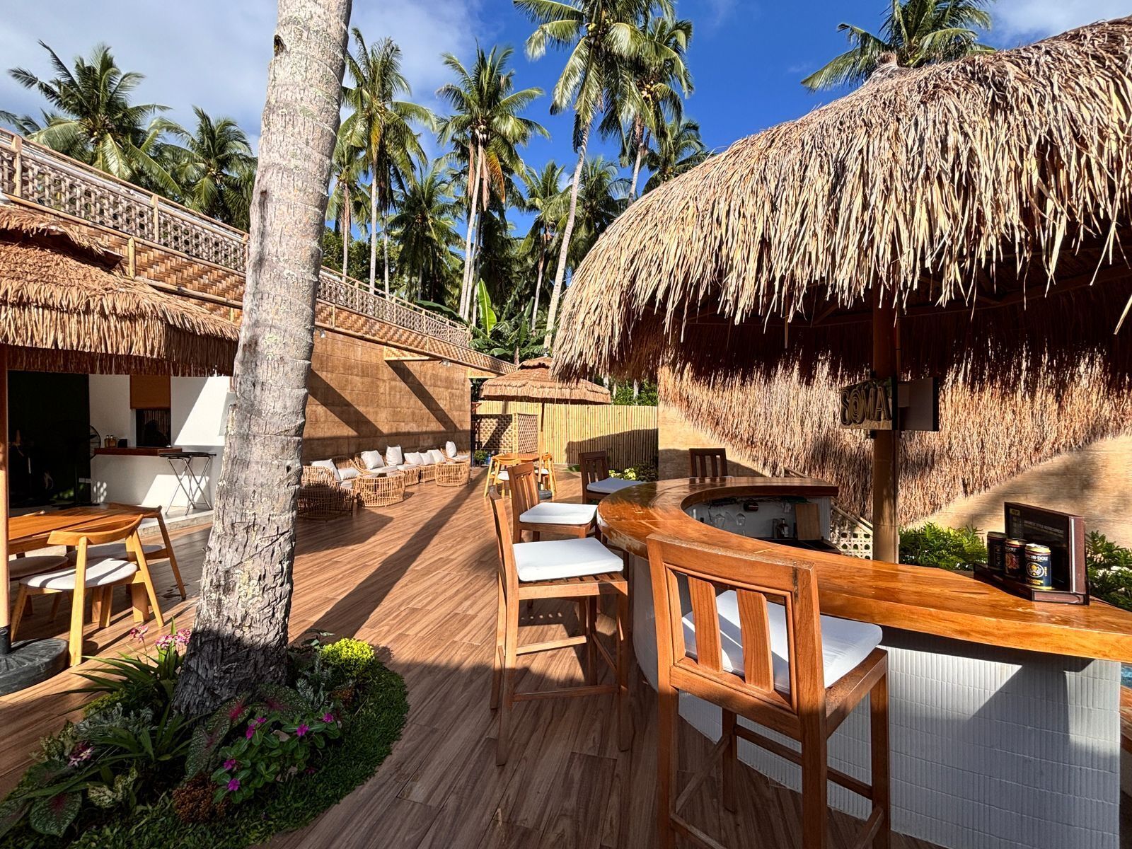 Un área de bar al aire libre con mobiliario de madera, techo de paja y vistas a palmeras.