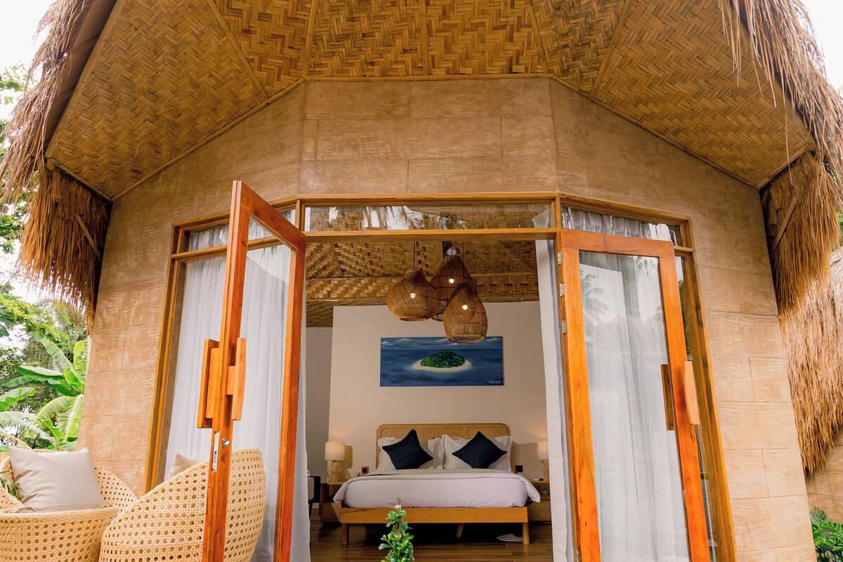 Uma cabana com telhado de palha, deck de madeira, cama com roupa de cama branca e móveis de vime.