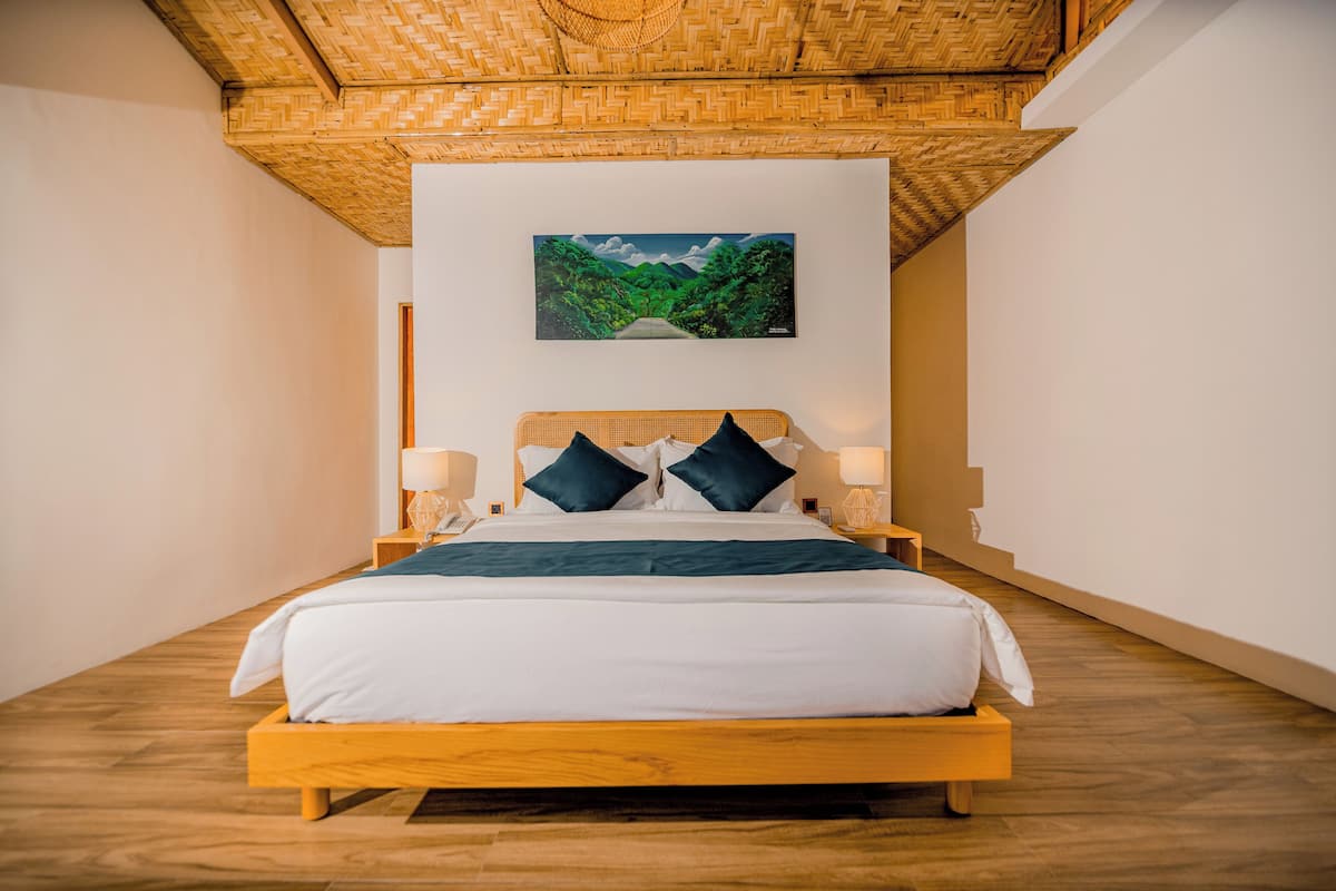 Quarto com cama de madeira, duas mesas de cabeceira com abajures, um quadro pendurado na parede representando uma floresta e teto com padrão em herringbone.