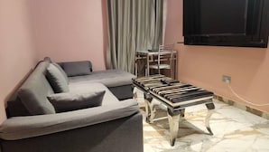 A living room with a grey sofa, a small table with books, and a TV mounted on the wall. - Triple Tee Luxury Hotel (Lagos)