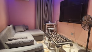 A living room with a grey sectional sofa, a small table with a chair, a standing fan, and a mounted TV. - Triple Tee Luxury Hotel (Lagos)
