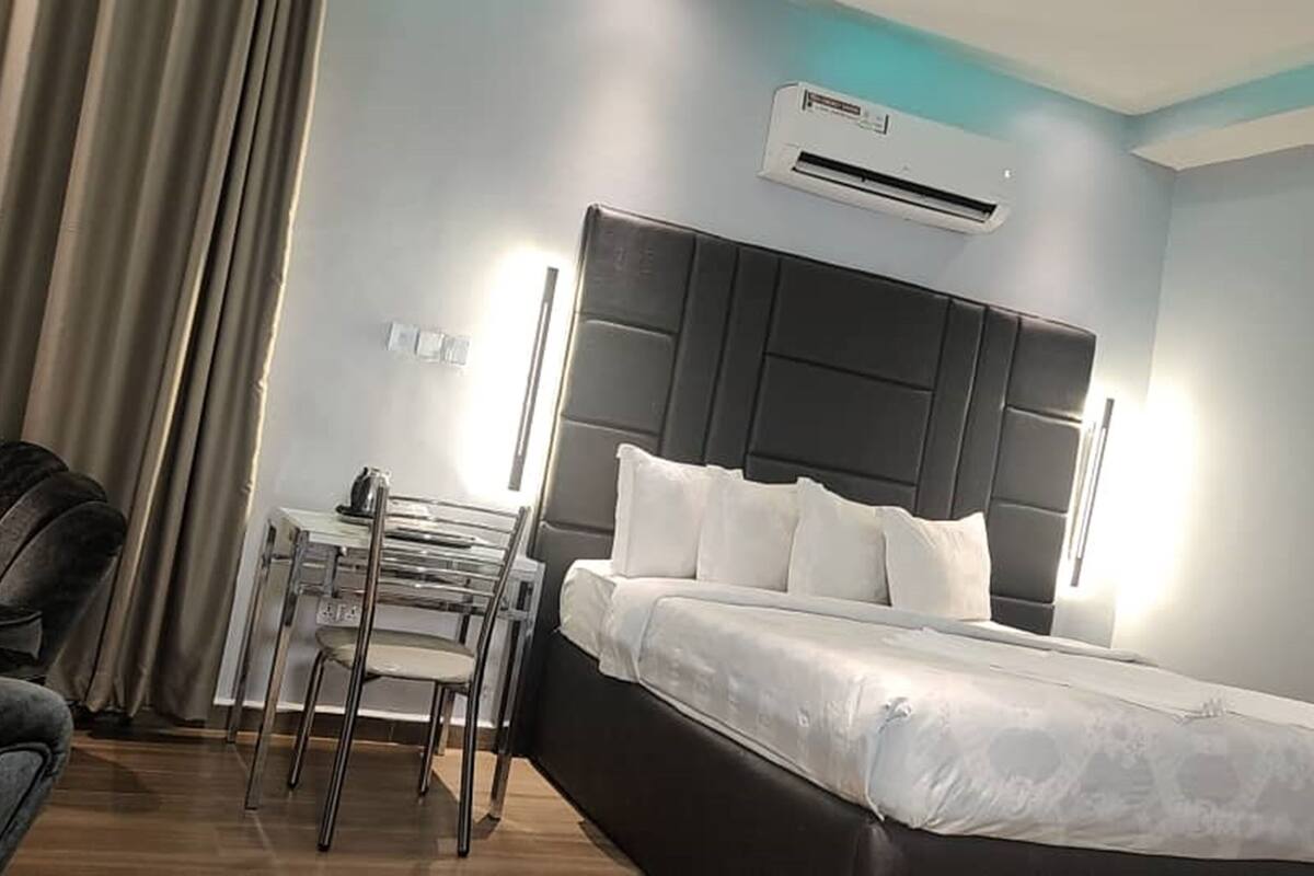 A hotel room with a bed, a desk, a chair, and an air conditioning unit.