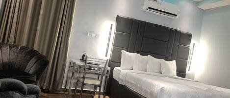 A hotel room with a bed, a desk, a chair, and an air conditioning unit.