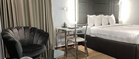 A modern hotel room with a large bed, a desk with a chair, and a dark armchair.