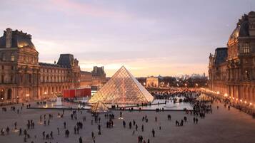 Une vaste place ouverte, dominée par une imposante structure pyramidale en verre au centre, et bordée d’édifices à l’architecture classique.