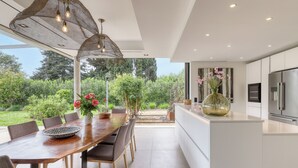 A modern kitchen with a large island, white cabinetry, and stainless steel appliances. There is a dining area with a wooden table and chairs, and a view of a lush garden.