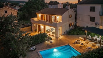 A two-story house with a swimming pool, outdoor seating, and a well-lit balcony.