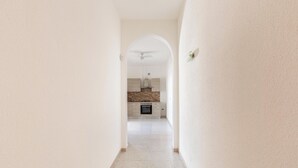 Un couloir mène à une cuisine au design moderne, dotée d’un ventilateur de plafond, d’un four encastré et d’un pare-feu dont la texture rappelle celle de la pierre.