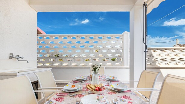 Une table à manger dressée avec des assiettes, des couverts et un vase de fleurs, entourée de chaises, avec une clôture blanche et un ciel dégagé en arrière-plan.