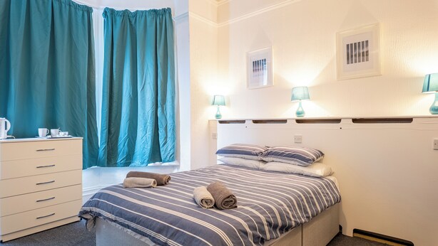 A bedroom with a bed, two bedside lamps, a window with curtains, a dresser, and a wall-mounted picture. - Pebbles Guest house (Southampton)