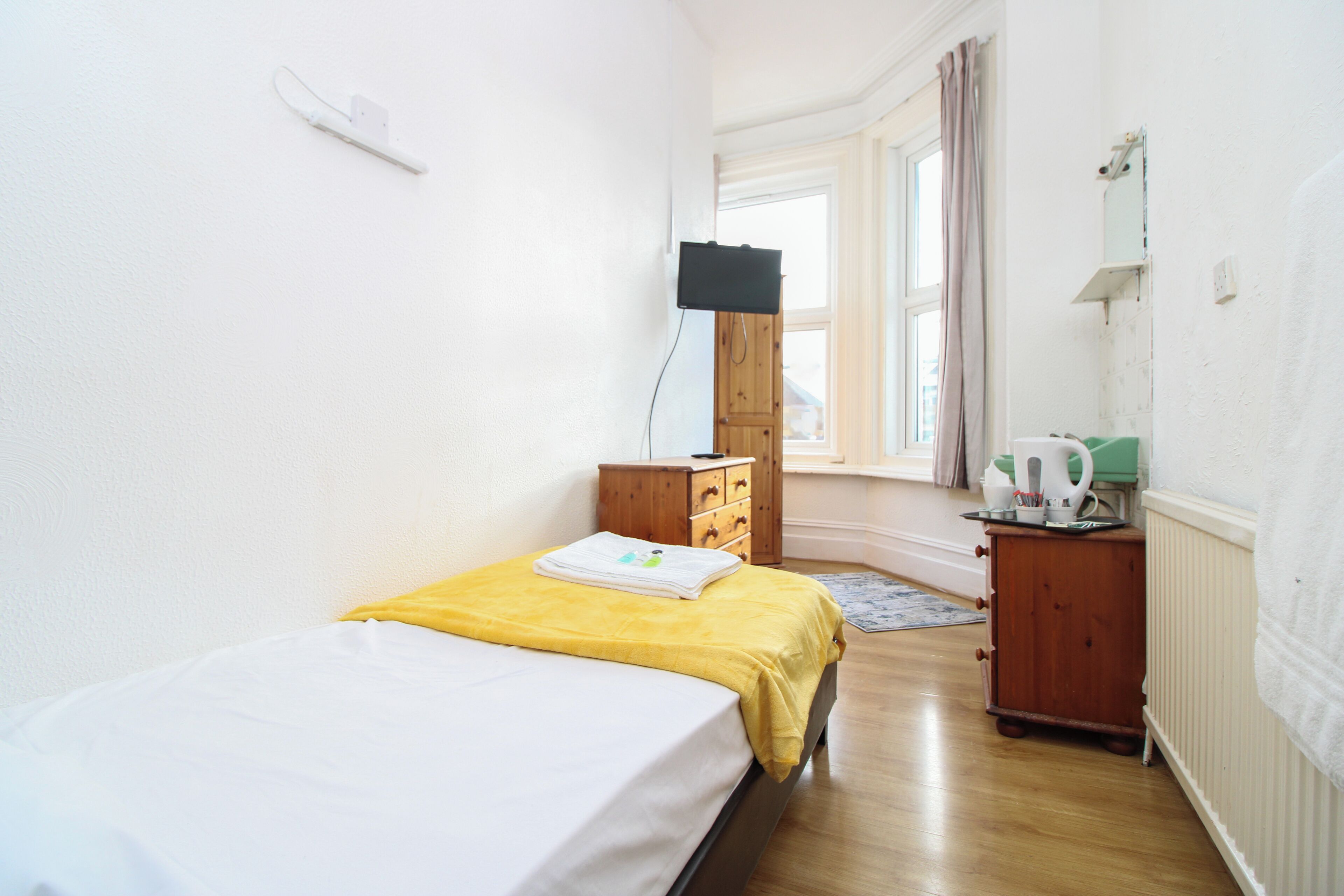 A small, bright room with a bed, a wooden dresser, a small table with a kettle, and a window with curtains.
