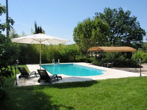 Piscina com guarda-sol branco e toldo de madeira, cercada por vegetação e árvores.
