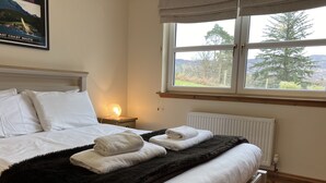 A neatly made bed with white linens and a black throw, a nightstand with a lamp, and a window with a view of trees and mountains. - Oiseval is a pet friendly cottage with magnificent views. (Fort Augustus)