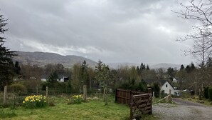 A rural landscape with a gravel path leading to a wooden gate, surrounded by greenery and a backdrop of mountains. - Oiseval is a pet friendly cottage with magnificent views. (Fort Augustus)