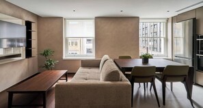 Un salon moderne comprenant un canapé, une table à manger, des chaises, un téléviseur fixé au mur et une grande fenêtre donnant sur la ville.