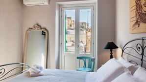 Hotelkamer met een bed, een grote spiegel, een raam met uitzicht op gebouwen, een stoel en een lamp.