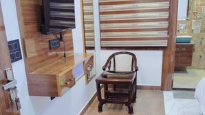 A room with a wooden floor, a wooden chair, a TV mounted on the wall, and a window with striped blinds. - Madhav Residency (Mathura)