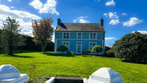 A two-story house with a blue exterior, a front lawn, and a set of stone steps leading to the front door. - Lisheenbawn Farmhouse Farranfore (Castleisland)