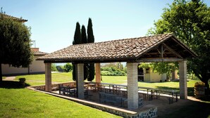 Un área de estar al aire libre cubierta con pilares de piedra y techo de baldosas, rodeada de césped y árboles.