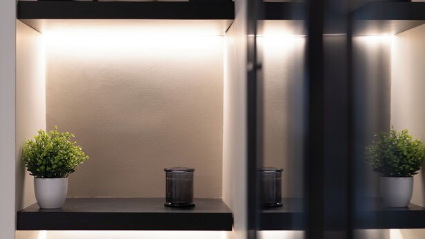Un intérieur moderne avec une étagère noire où sont posées des cartes encadrées, un vase blanc contenant une plante et un chandelier noir.