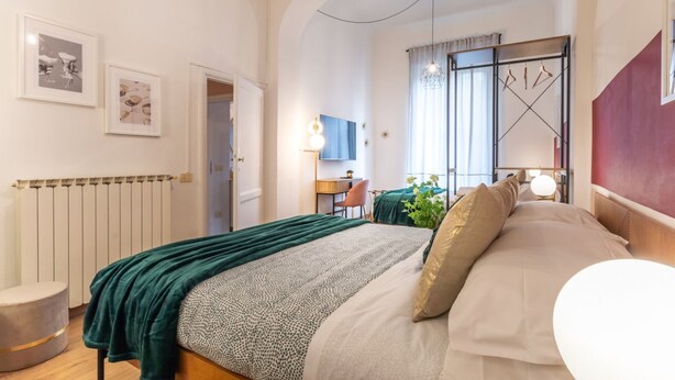 A modern bedroom with a large bed, a desk, and a chair. There is a radiator, a plant, and a window with curtains. - Wine Apartments Florence Prosecco (Firenze)