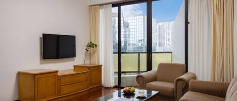 A modern living room with a flat-screen TV, wooden furniture, a coffee table with a book and fruit, and a large window with curtains.