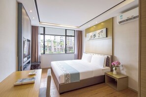 A modern hotel room with a large bed, a desk with a chair, a small table with a vase of flowers, and a window with a view of buildings and palm trees.