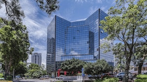 Un bâtiment moderne en verre, avec une entrée située à l’angle, entouré d’arbres et de voitures garées.