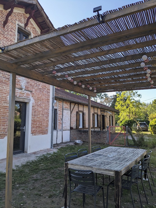 Un espacio de estar al aire libre cubierto, con una mesa y sillas, un edificio de ladrillo con ventanas y una pérgola de madera.