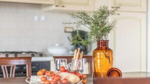Une cuisine avec une table mise pour le repas, agrémentée d’un vase contenant des feuilles vertes, d’une bouteille et d’une assiette avec des tomates et du pain.
