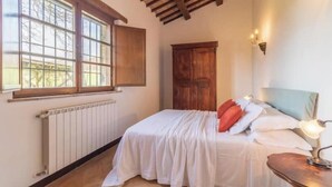 Une chambre avec un plafond en bois, un radiateur, un lit avec une literie blanche et des oreillers rouges, une table de chevet avec une lampe et une armoire en bois.
