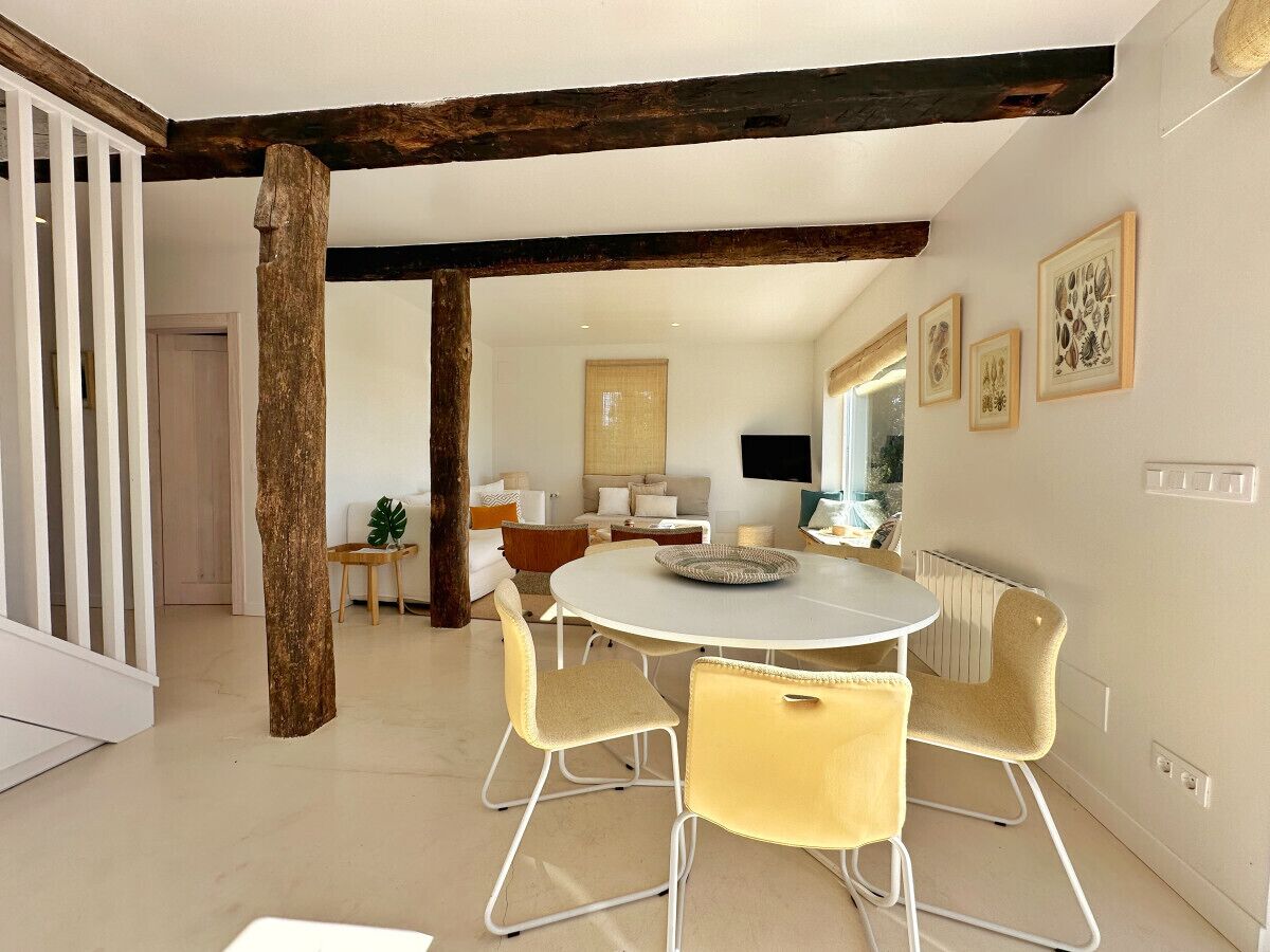 Ein Essbereich mit einem runden Tisch, gelben Stühlen und einer weißen Tischdecke. Im Hintergrund sind Holzbalken an der Decke und eine weiße Geländer zu sehen.