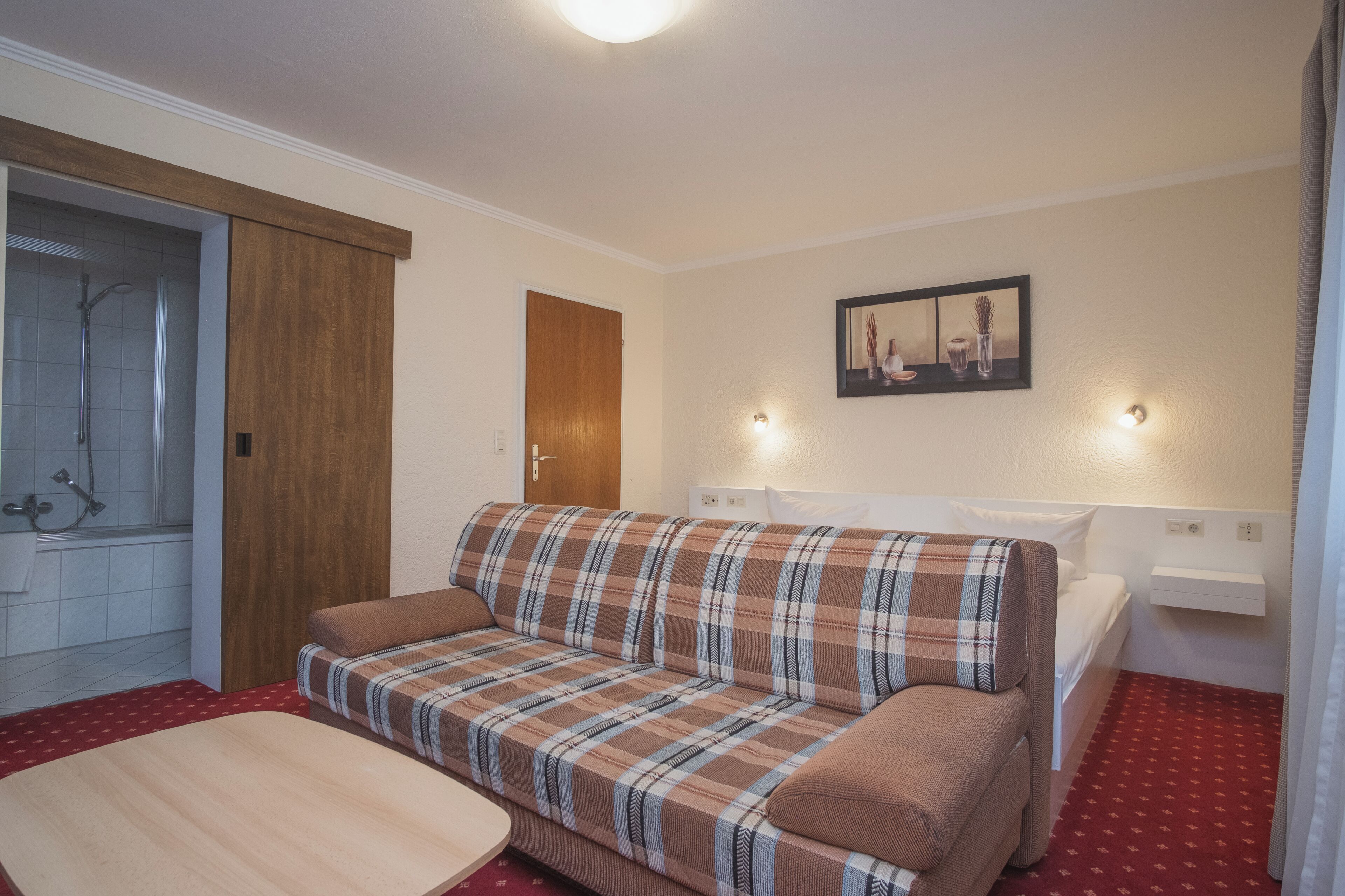 A room with a plaid sofa, a wooden door, a shower, and a small bedside table.