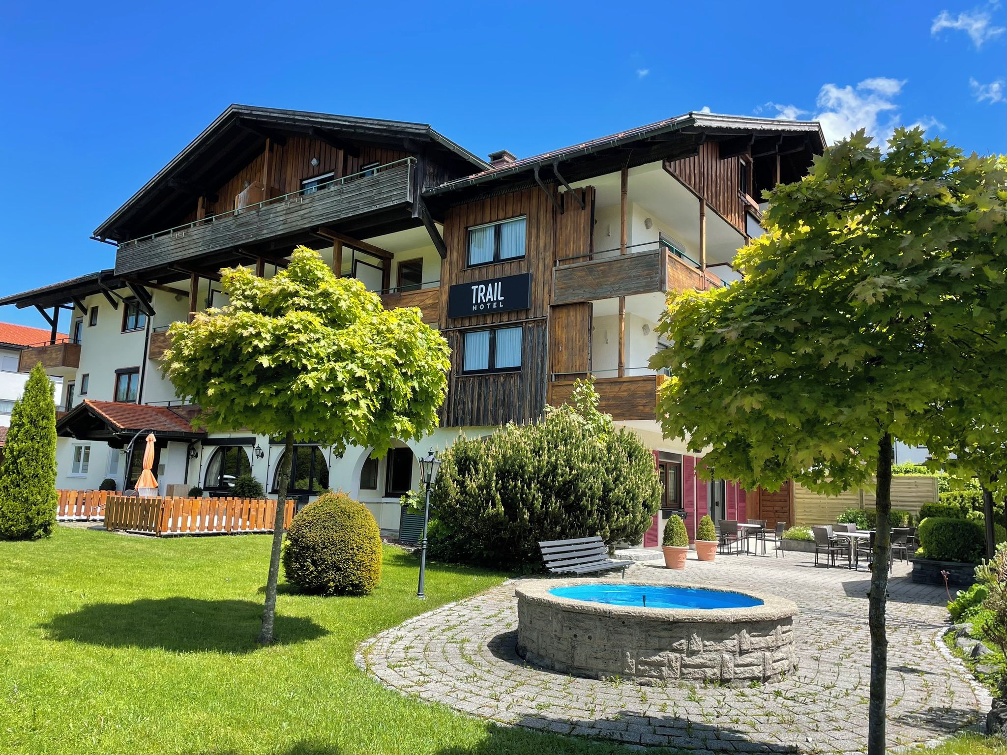Un hôtel avec une façade en bois, une enseigne indiquant « TRAIL HOTEL » et une fontaine circulaire en pierre dans la cour.