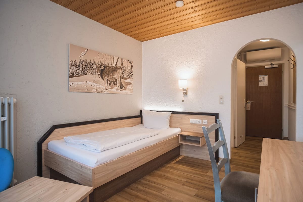 A room with a wooden bed, a desk, a chair, a wall-mounted light, and a door.