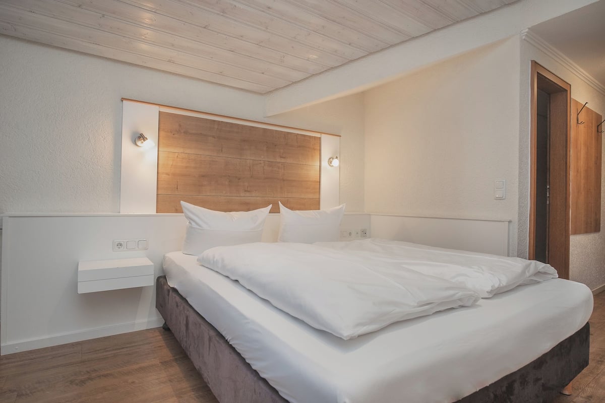 A bedroom with a wooden headboard, white bedding, a bedside table, and a wall-mounted light fixture.